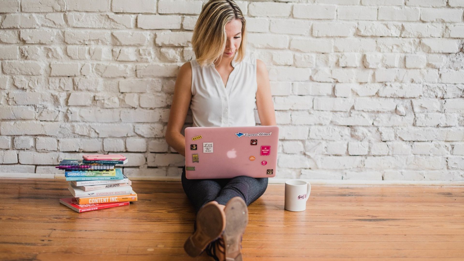 A girl sitting down on the floor, back towards a white wall and legs straight in front of her. She has a computer on her lap, books to her right and a coffee mug to the left. The image represents the AI vs. Human Generated Content drama.