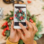 A picture of someone taking a picture of a holiday wreath on a table along with other holiday decorations. Probably to use it as holiday content for socials.