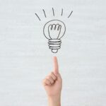 A hand pointing to a drawing on a lightbulb on a gray background symbolizing motivation and inspiration.