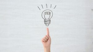 A hand pointing to a drawing on a lightbulb on a gray background symbolizing motivation and inspiration.