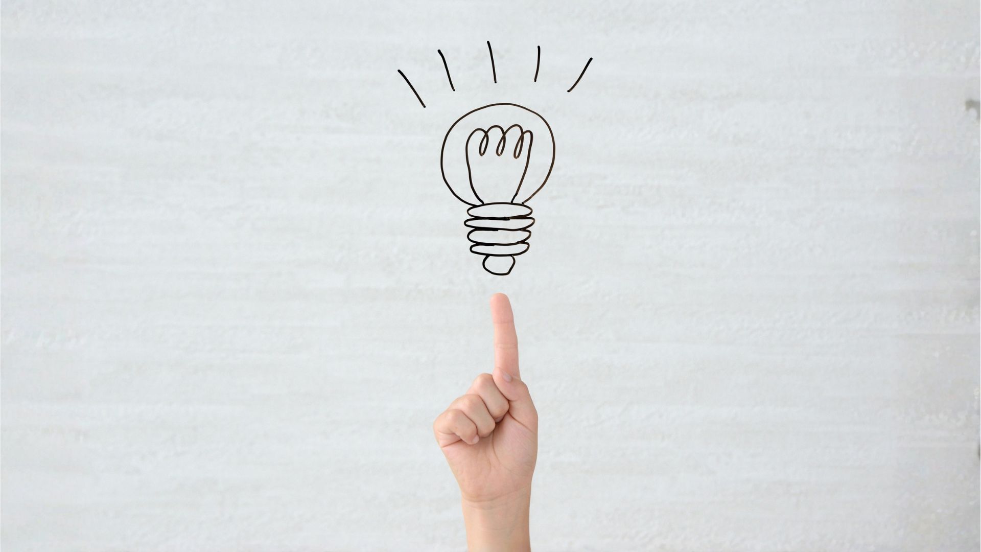 A hand pointing to a drawing on a lightbulb on a gray background symbolizing motivation and inspiration.