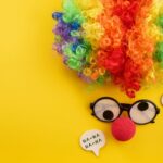 A clown wig with silly glasses and a red nose on a yellow background to represent April Fools day.