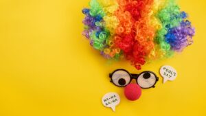 A clown wig with silly glasses and a red nose on a yellow background to represent April Fools day.