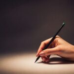 The hand of a woman, holding a pencil as if it is about to start writing. Is the hand real? Is it an allegory to AI vs. Human Written Content?